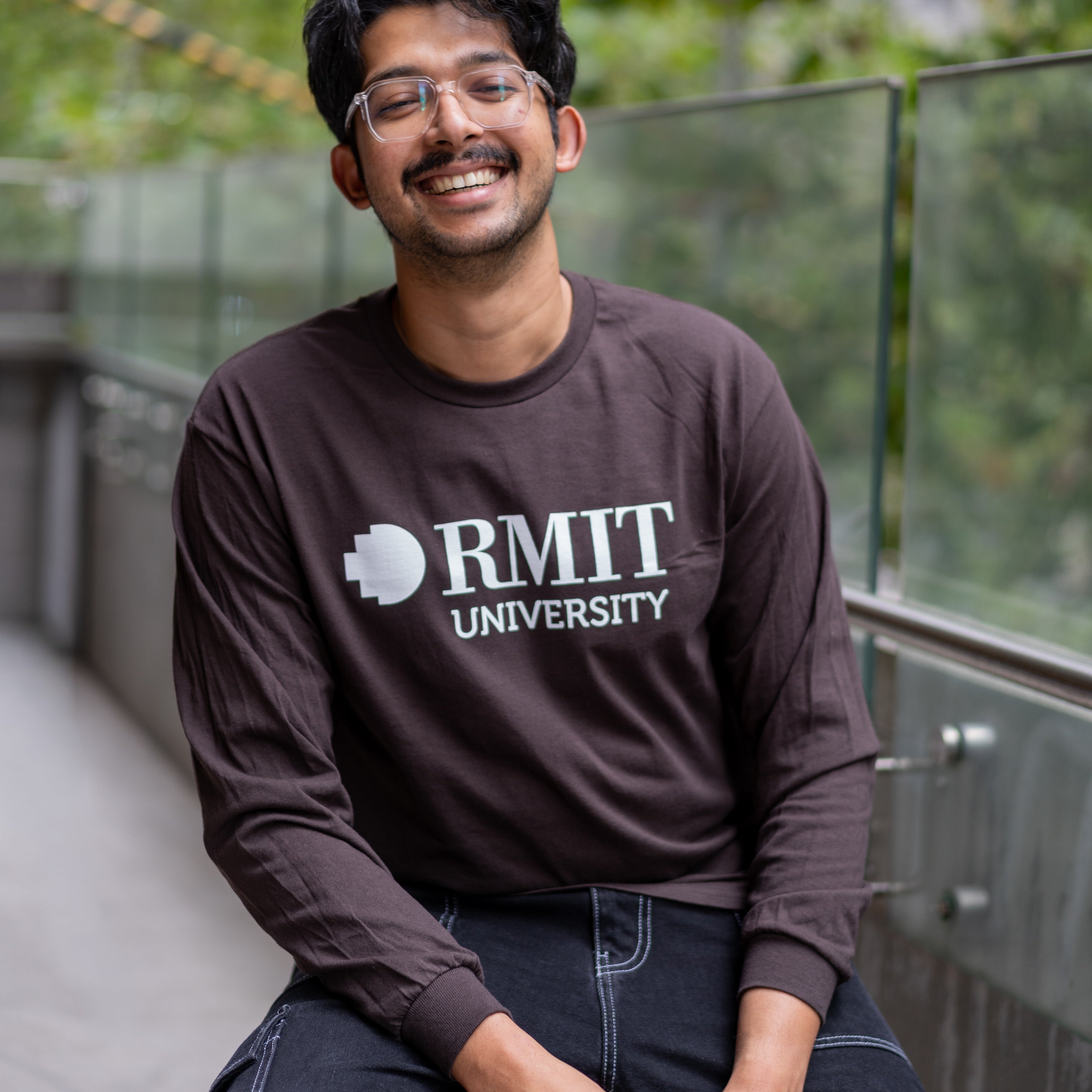 Student wearing RMIT Dawson long sleeve tee in chocolate brown with white RMIT University logo, smiling while seated outdoors on campus.