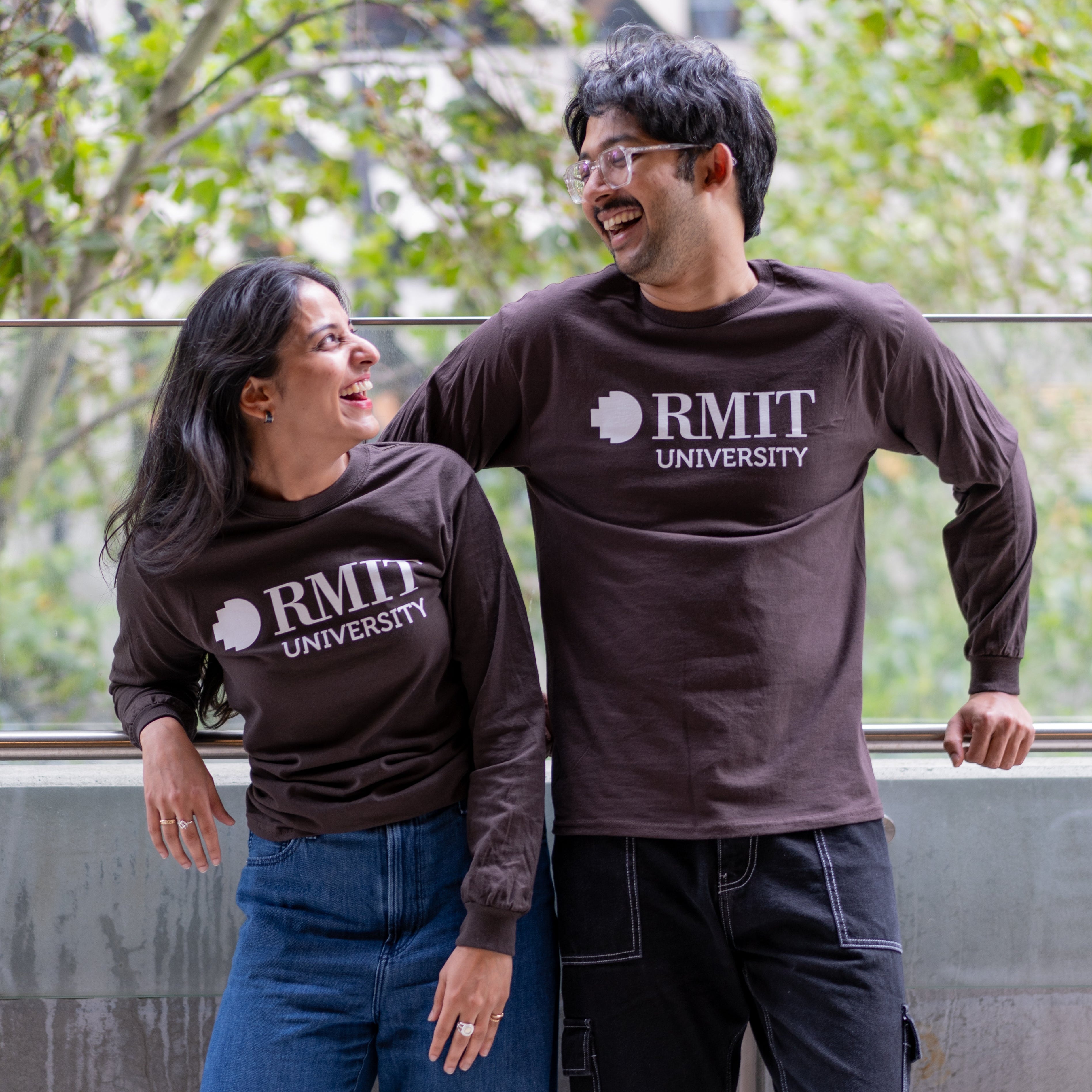 Two students wearing RMIT Dawson long sleeve tees in chocolate brown with white RMIT University logo, smiling at each other outdoors on campus.