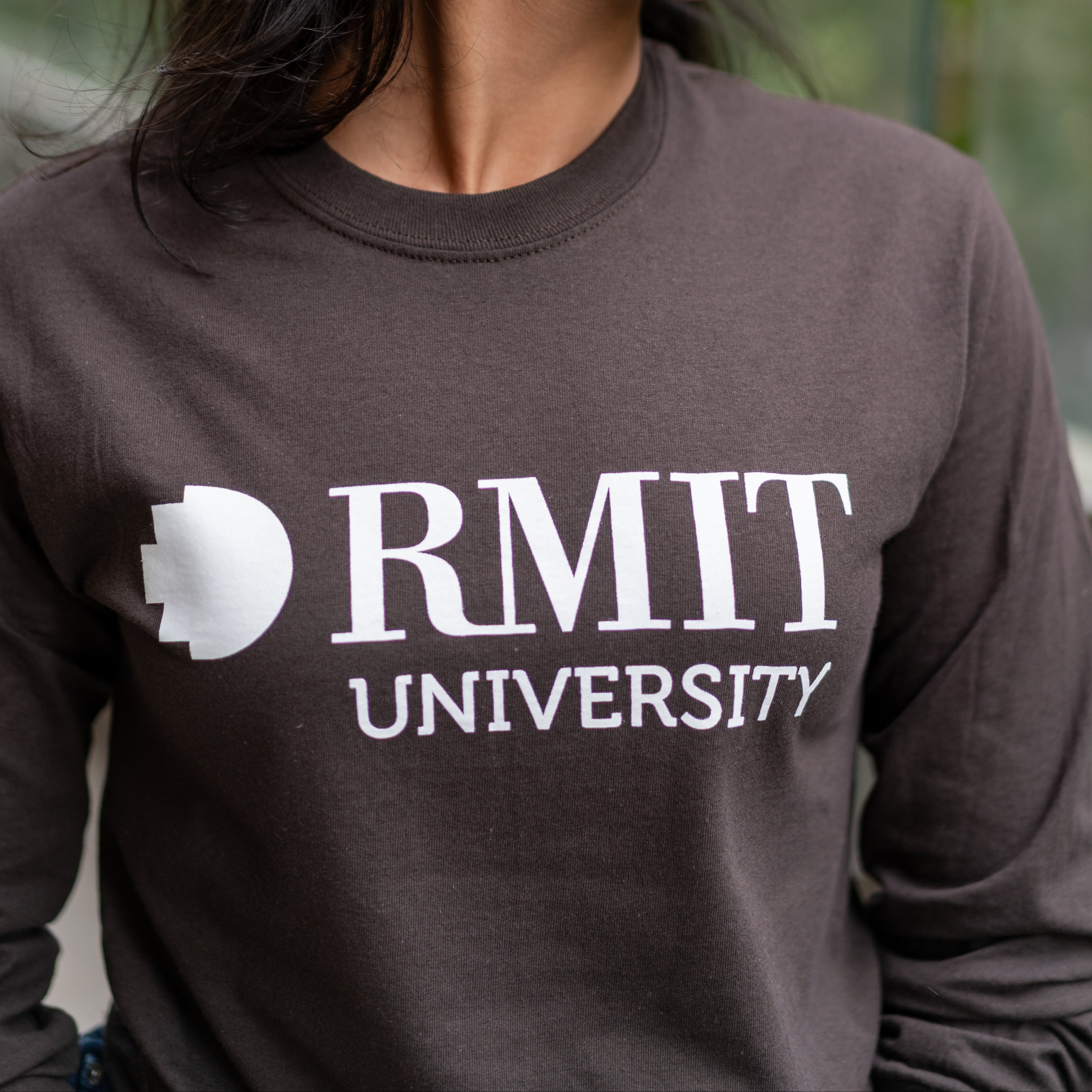 Close-up of student wearing RMIT Dawson long sleeve tee in chocolate brown with white RMIT University logo, looking to the side on campus.