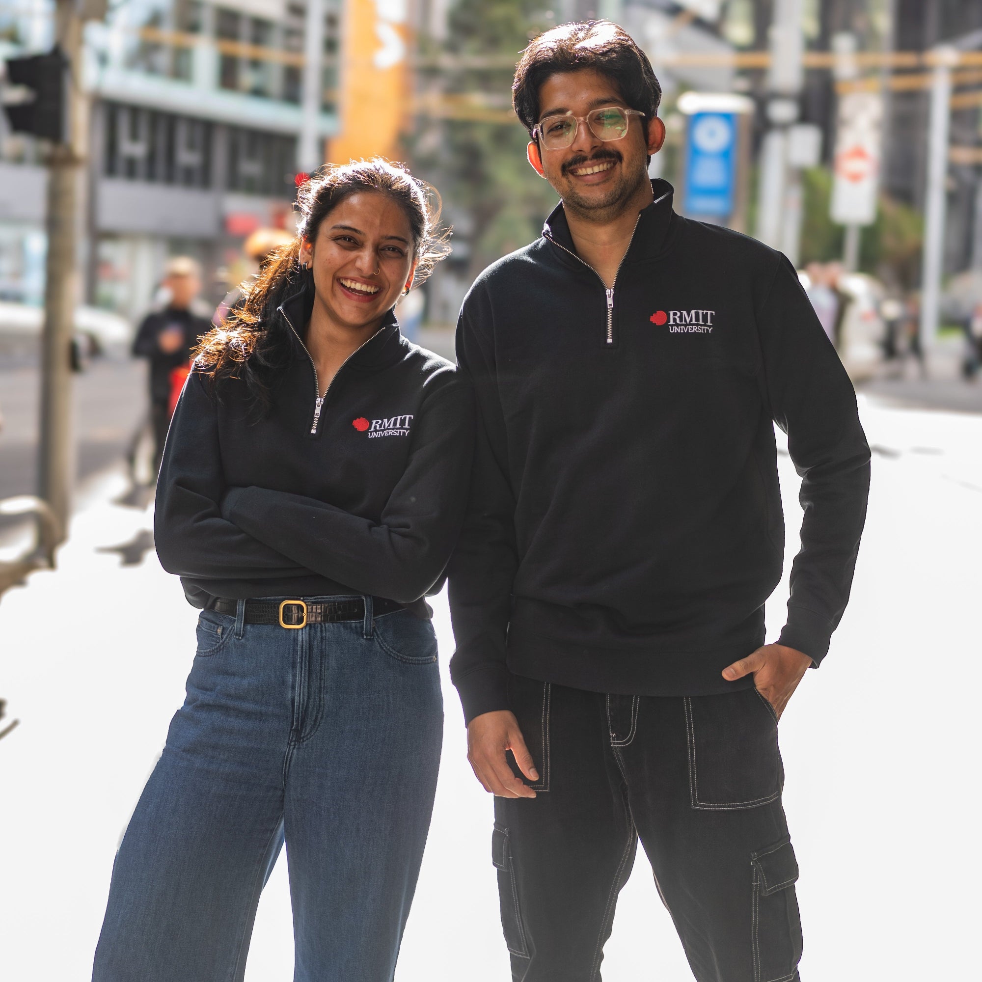 Two students wearing RMIT Essential half zip jumper in navy with embroidered RMIT University logo, standing together outdoors in the city and smiling.