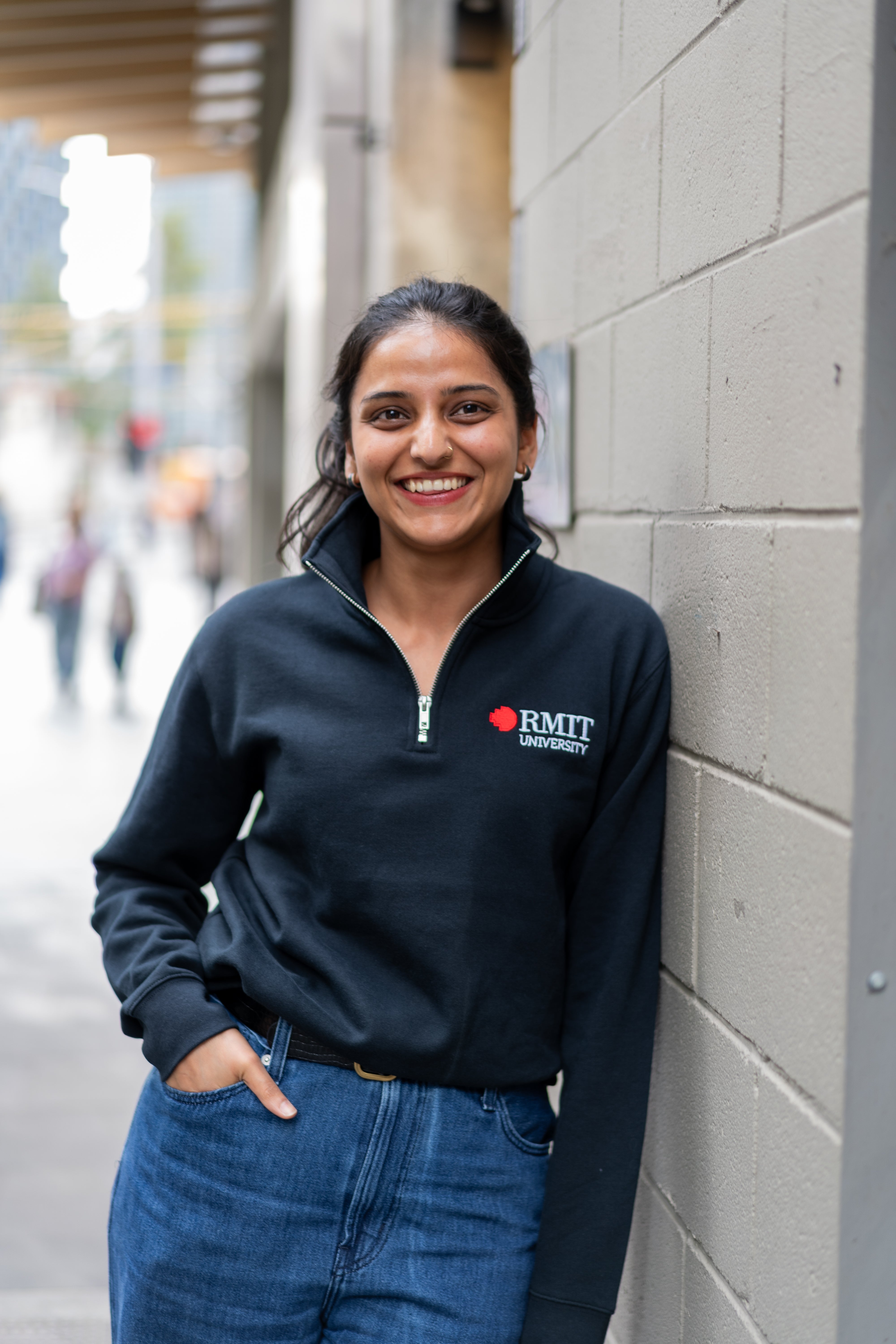 Student wearing RMIT Essential half zip jumper in navy with embroidered RMIT University logo, smiling while standing against a wall on campus.