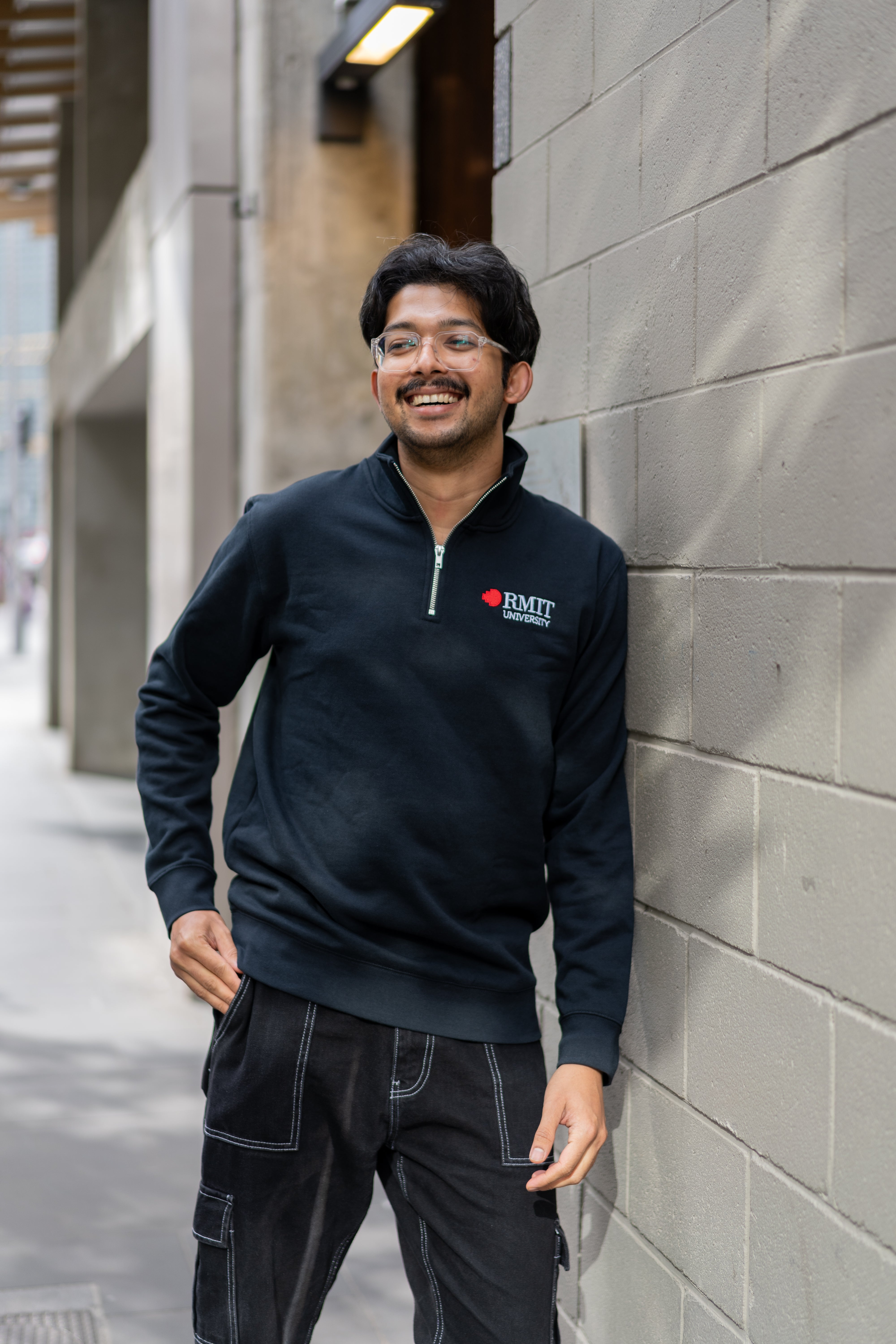 Student wearing RMIT Essential half zip jumper in navy with embroidered RMIT University logo, standing outdoors against a wall on campus and smiling.