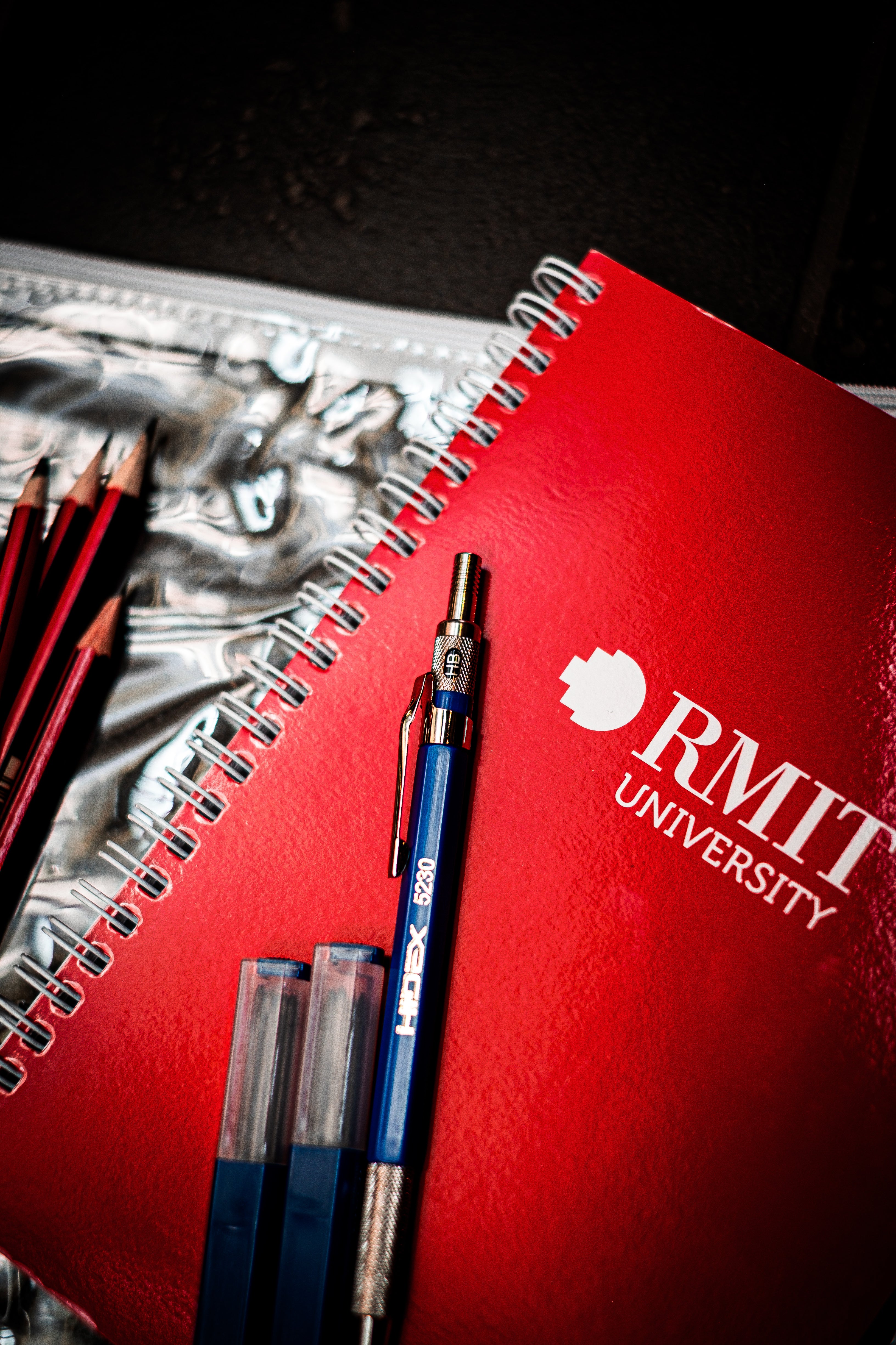 Close-up of RMIT Store's Interior Design Kit components: RMIT branded visual diary, Hidex mechanical pencil, and pencil leads arranged in a plastic pouch.