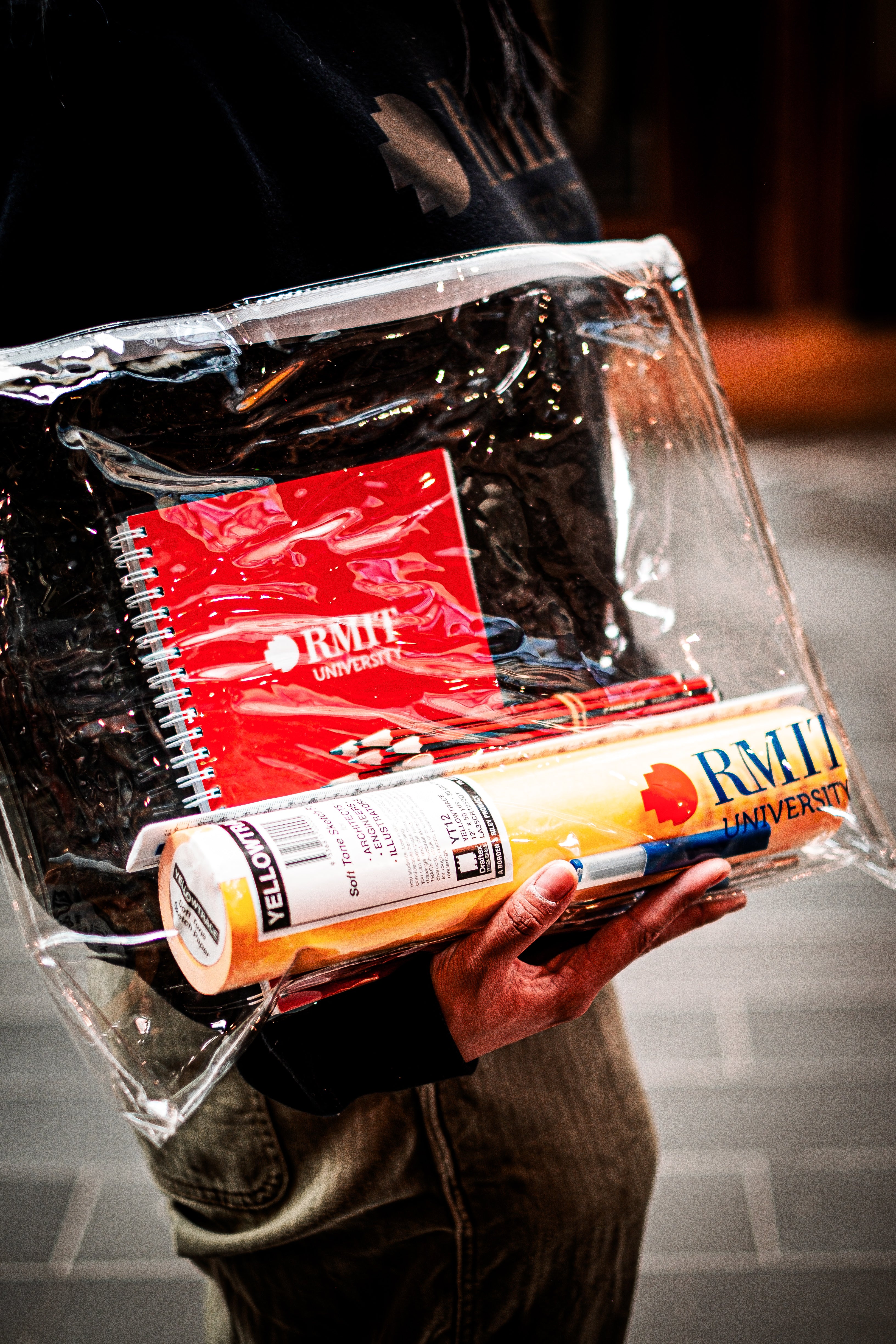 Hand holding RMIT Store's Interior Design and Decoration Kit, highlighting the RMIT visual diary, Yellowtrace sketch paper, assorted pencils contained in a transparent pouch and pencil leads contained in a clear pouch.