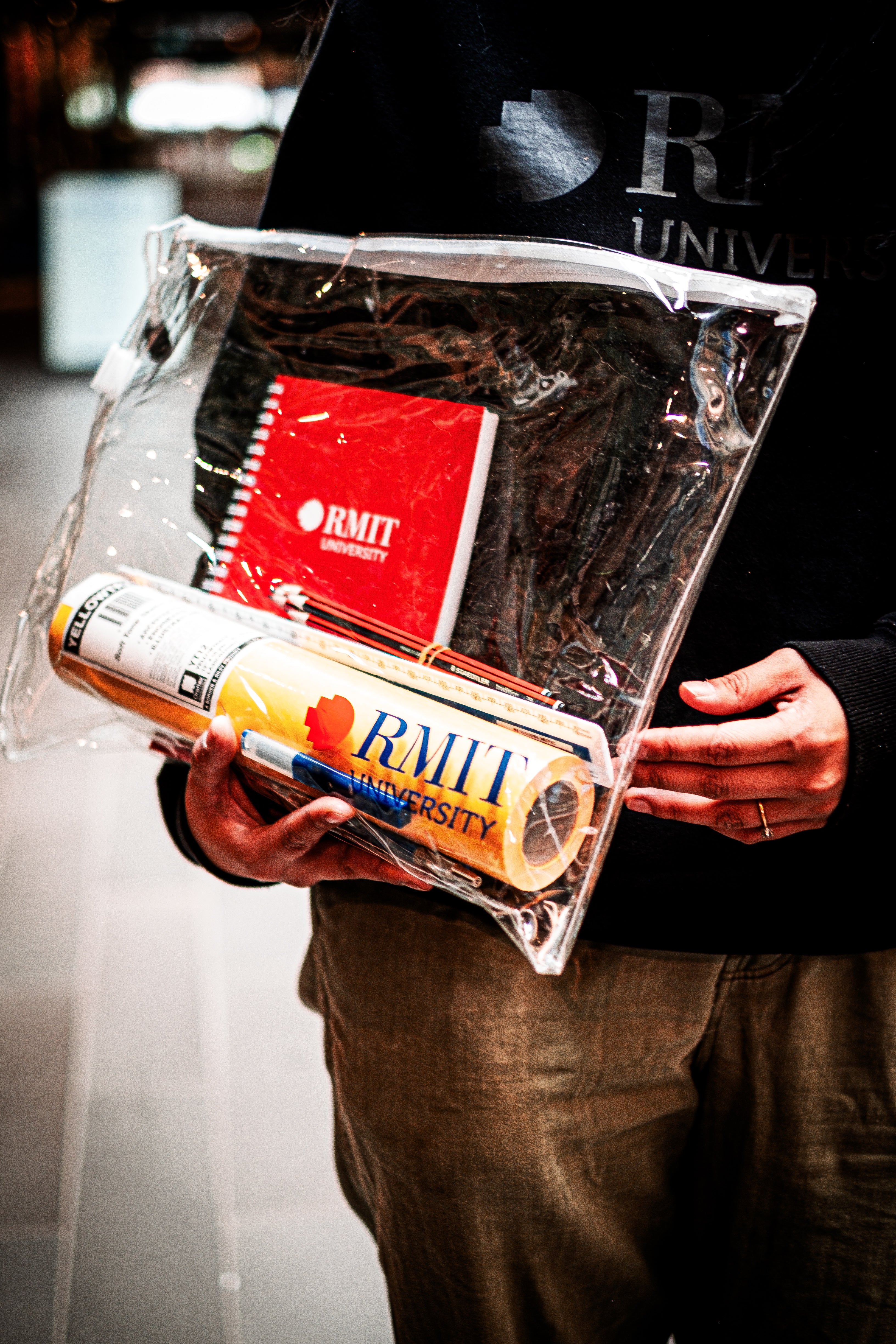 Hand holding RMIT Store's Interior Design and Decoration Kit, featuring an RMIT branded A5 visual diary, Yellowtrace sketch paper, and assorted pencils in a clear plastic pouch.