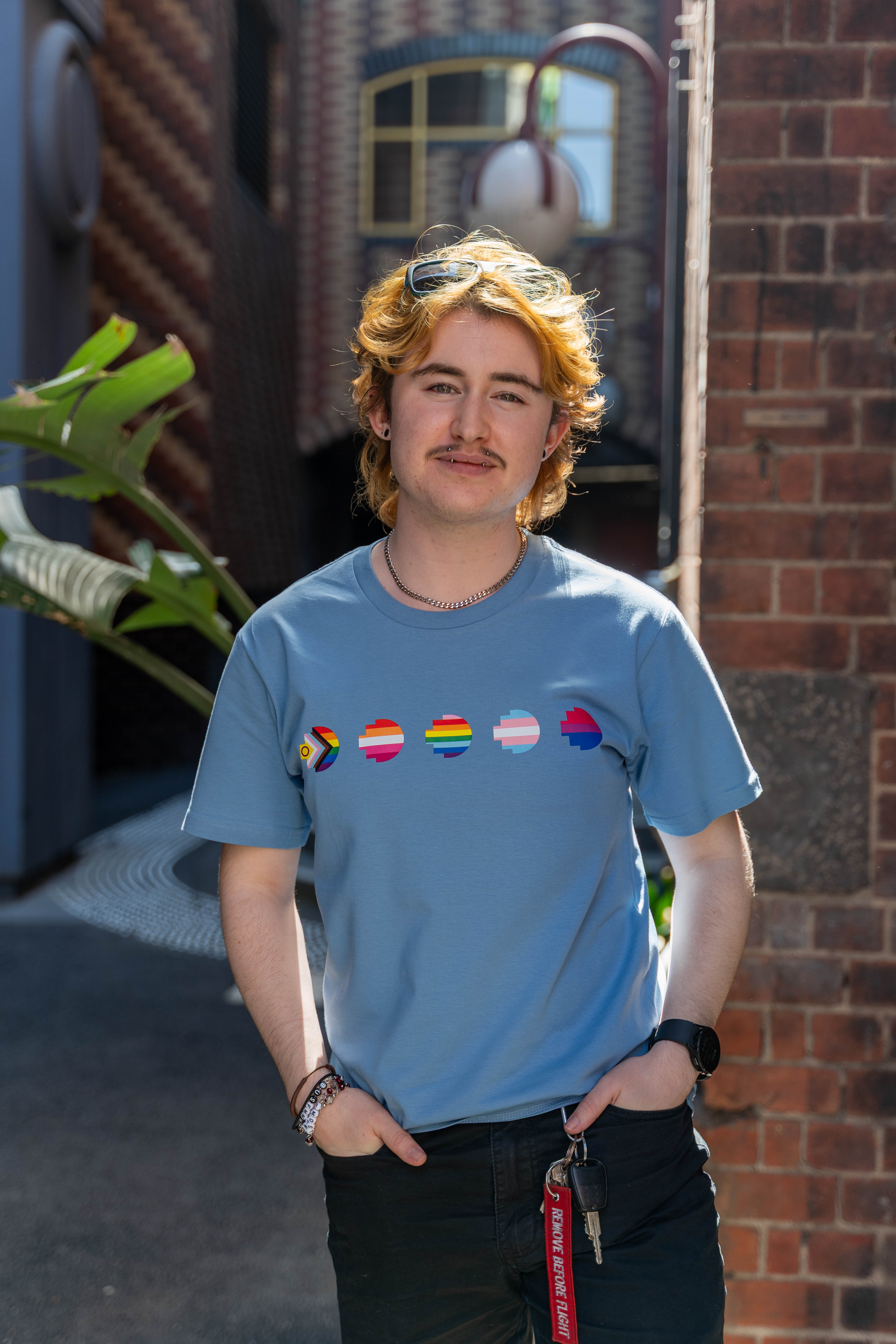 Modelled front view of the limited edition blue Pride Unity Tee from RMIT Store, displaying pride flags in a row across the chest in a contemporary urban backdrop.
