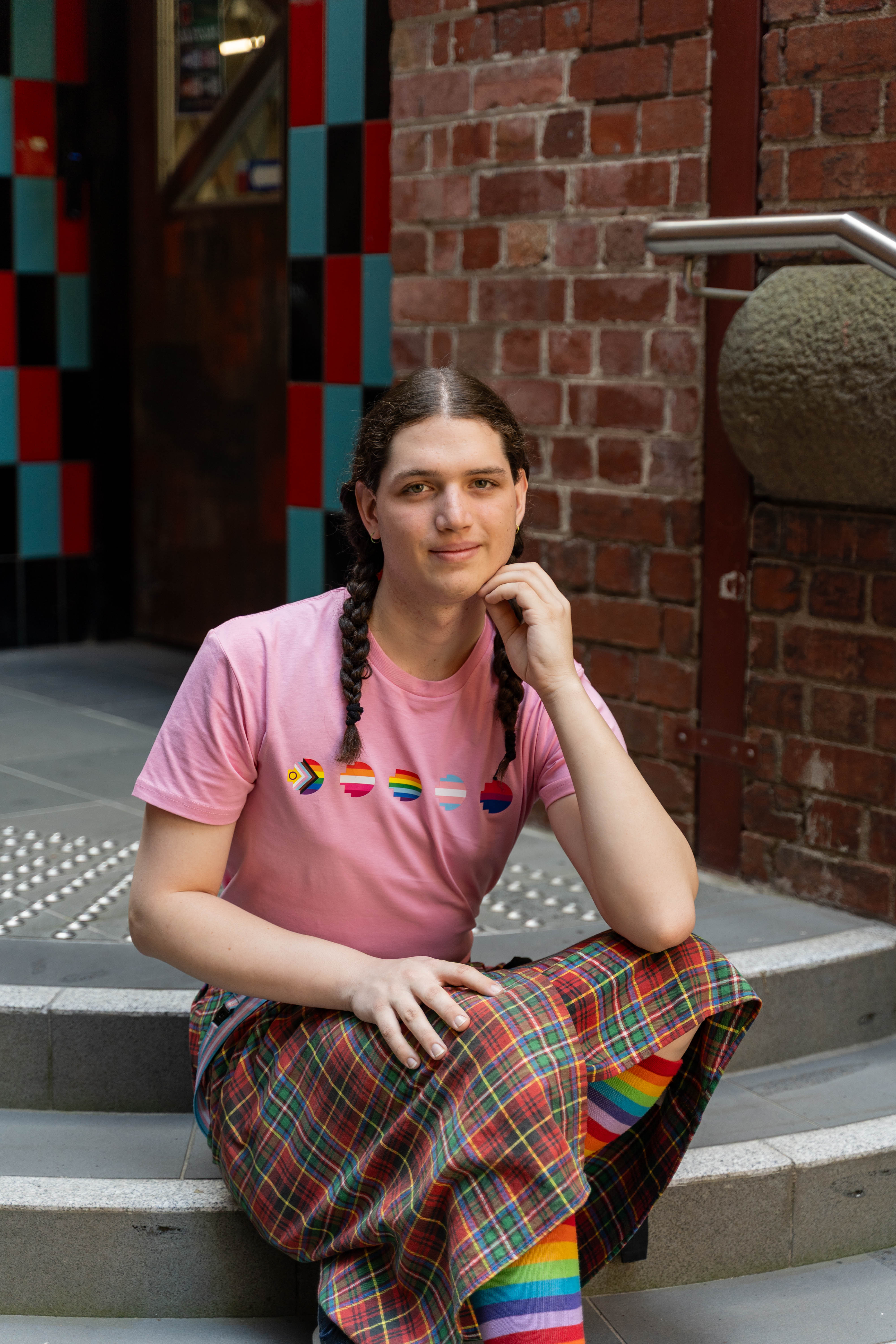 Model wearing the limited edition pink Pride Unity Tee from RMIT Store with pride flag designs, seated with rainbow socks in a colourful plaid skirt.