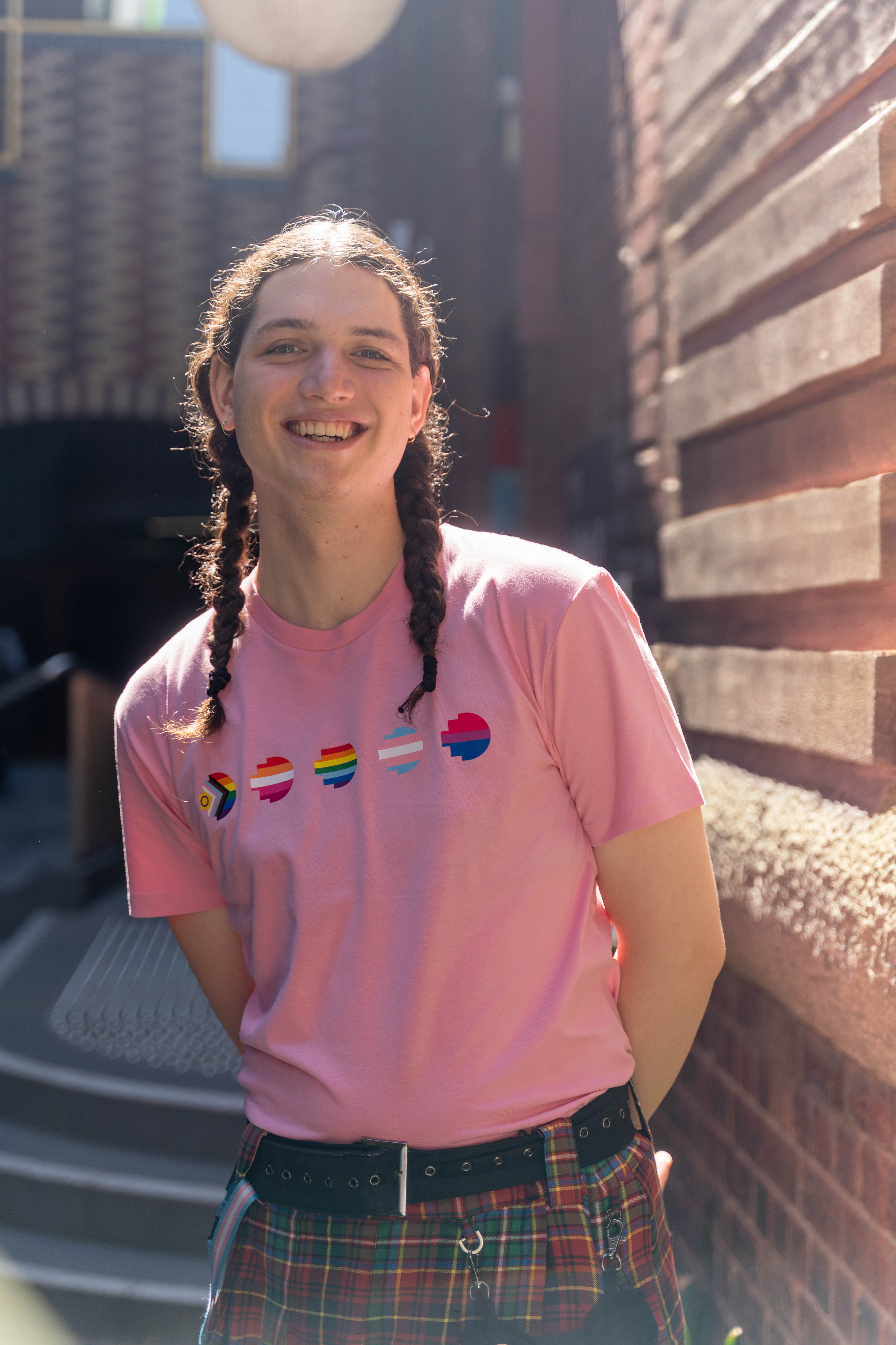 Model in a pink Pride Unity Tee from RMIT Store, featuring pride flag designs on the chest, styled with a plaid skirt in an urban alleyway.