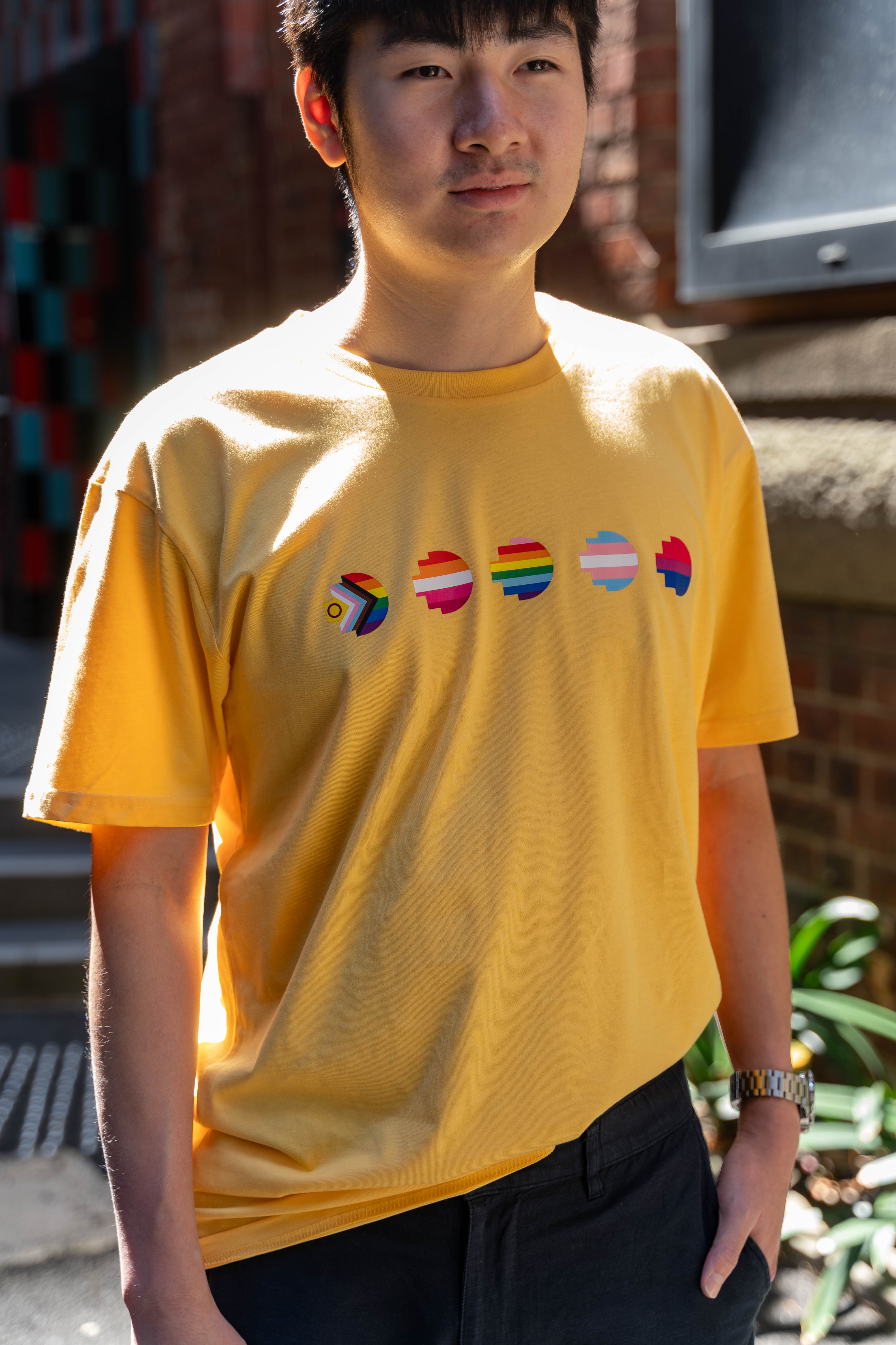 Front view of model wearing the yellow limited edition Pride Unity Tee from RMIT Store, complete with pride flag graphics set against a city backdrop.