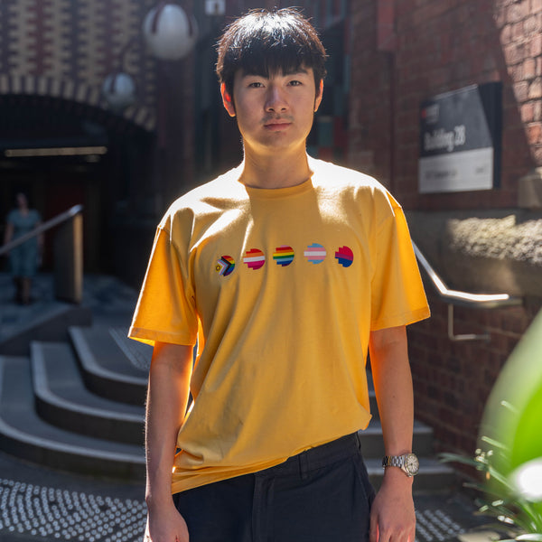 Model wearing the limited edition yellow Pride Unity Tee from RMIT Store, featuring colourful pride flag designs across the chest in a sunny urban setting.