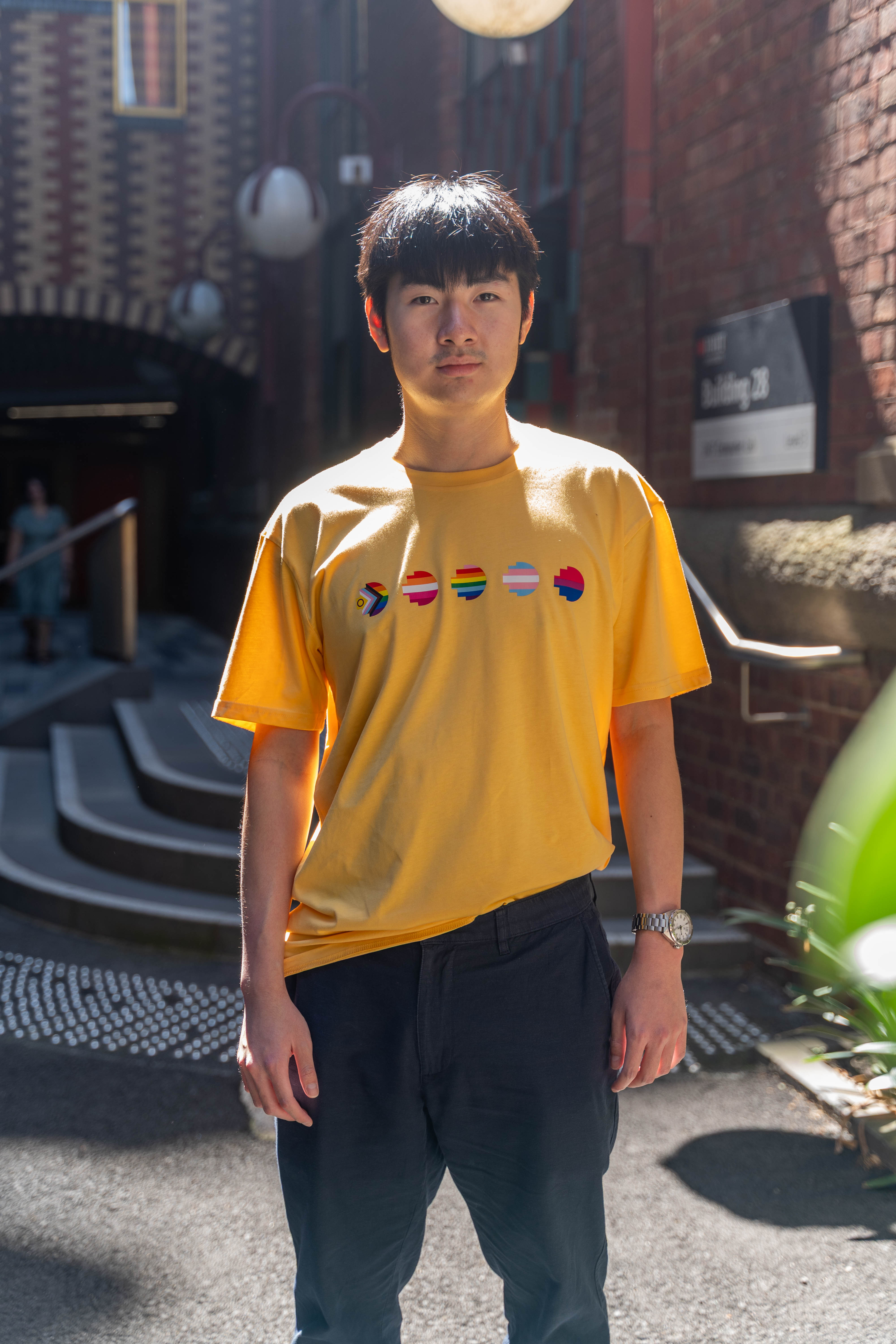 Model wearing the limited edition yellow Pride Unity Tee from RMIT Store, featuring colourful pride flag designs across the chest in a sunny urban setting.