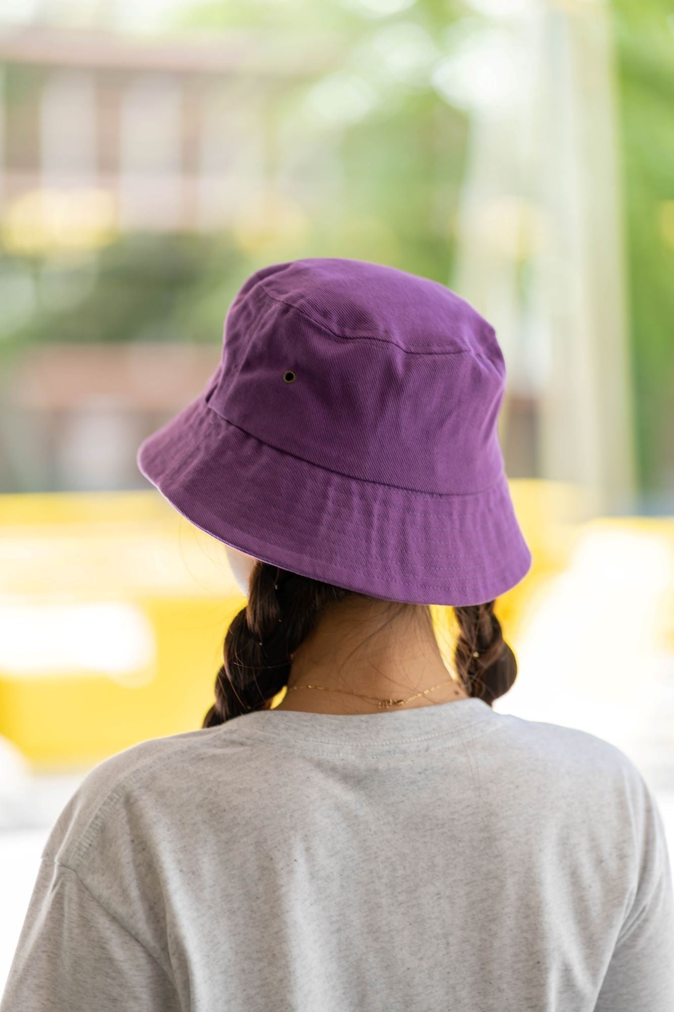 Rear view of a model wearing a purple RUSU bucket hat, highlighting its simple and vibrant design against a blurred outdoor background. The outfit includes a casual grey T-shirt that complements the hat's style, all part of the RMIT Store collection.