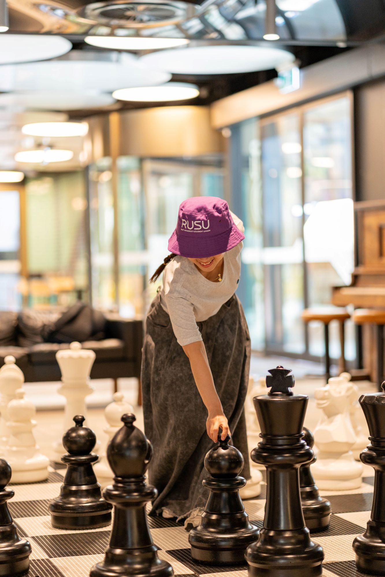 Model in a purple RUSU bucket hat with RMIT University Student Union branding engaged in a chess game with oversized black and white pieces. The model wears a grey long-sleeve top and a grey skirt, illustrating the hat’s contemporary appeal for student life.