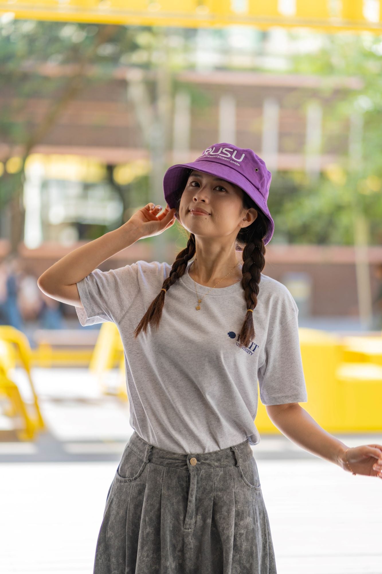 Model wearing a purple RUSU bucket hat with RMIT University Student Union branding, standing outdoors in a casual grey T-shirt and a grey pleated skirt, holding the brim of the hat. RMIT Store product showcasing a relaxed and stylish design suitable for everyday wear.