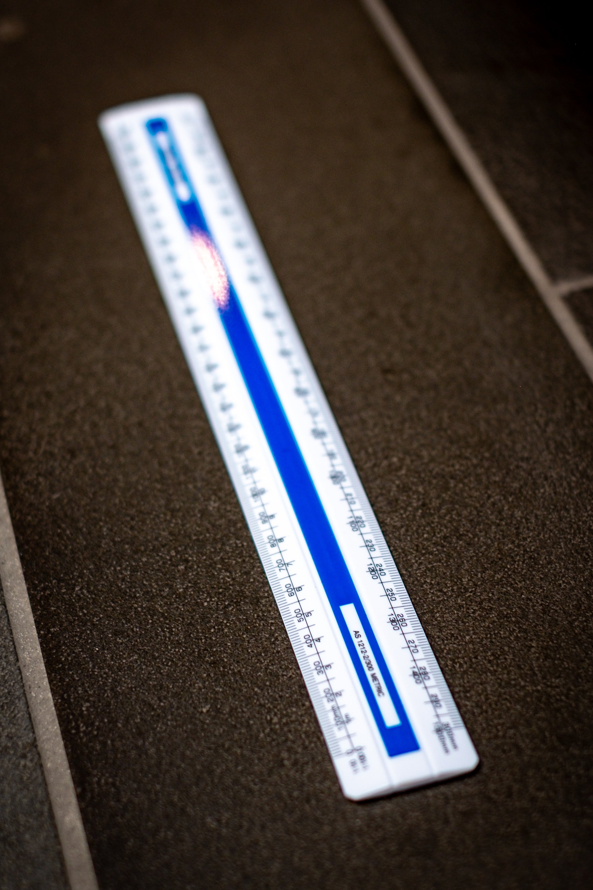 Angled side view of the Scale Ruler M62 from the RMIT Store, set against a dark textured background, highlighting its precision in metric scale markings.