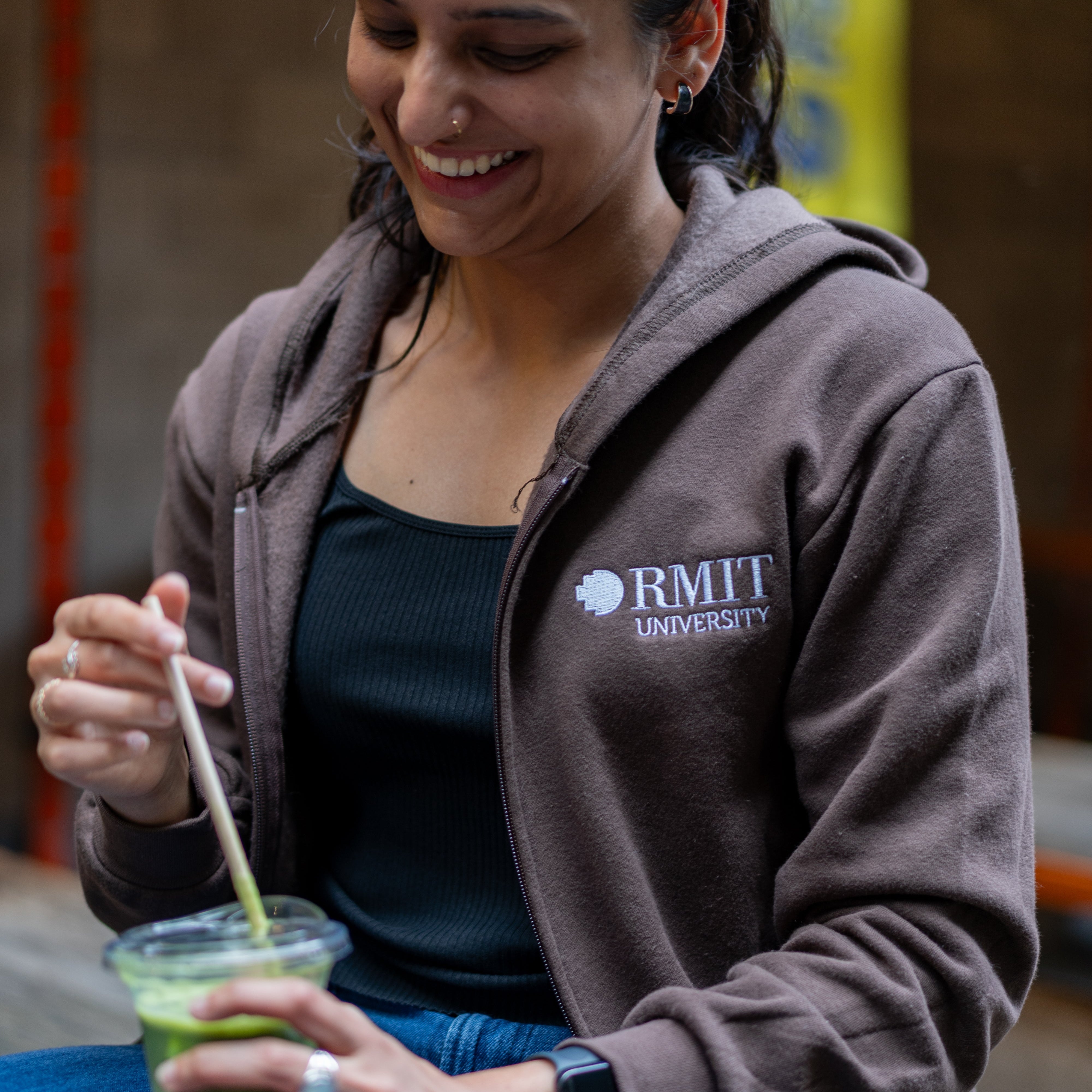 Student wearing RMIT University zip jacket in chocolate brown with white RMIT logo, sitting on campus and holding a drink.