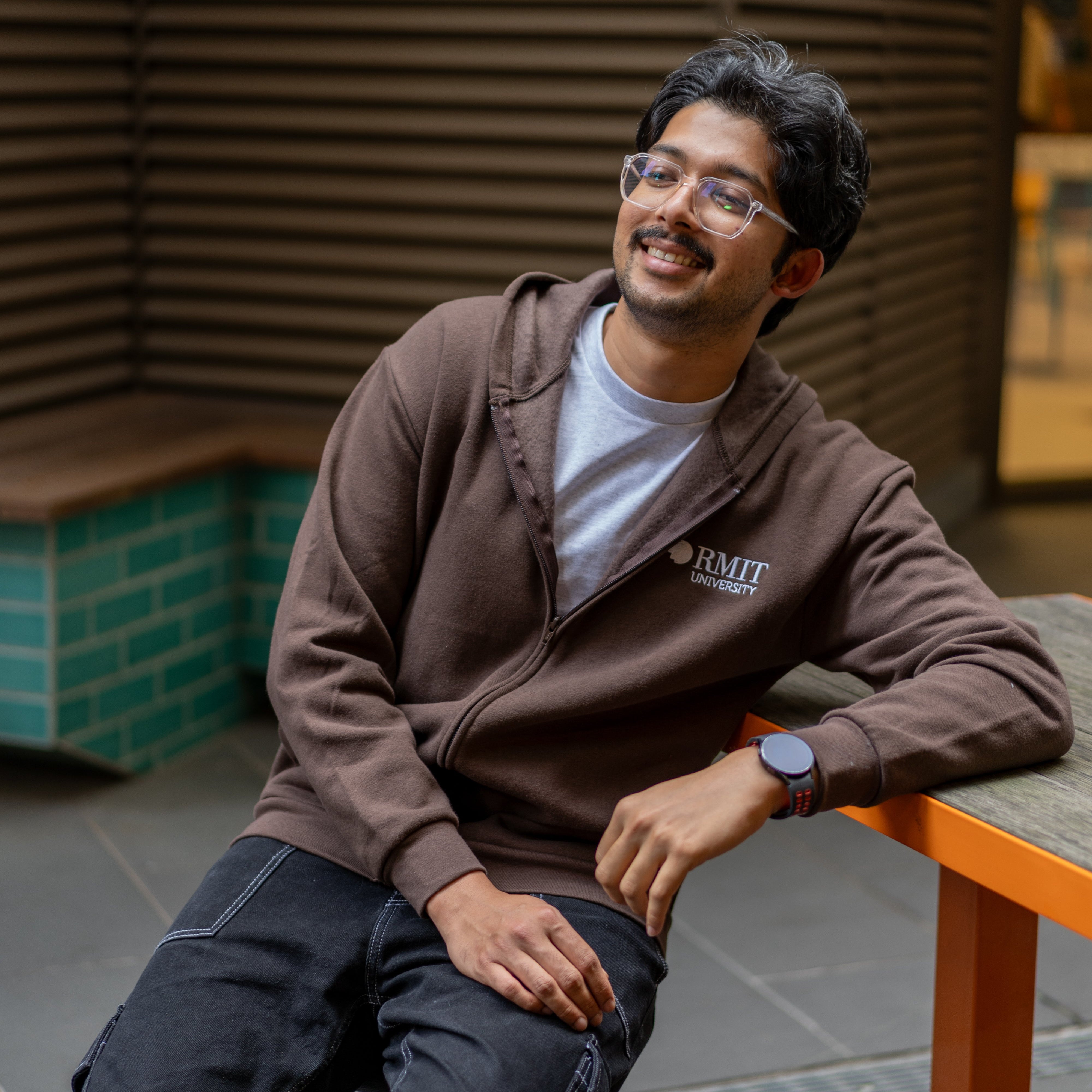 Student wearing RMIT University zip jacket in chocolate brown with white RMIT logo, seated and smiling on campus.