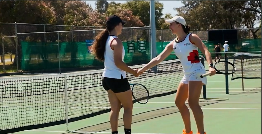Two female RMIT Redbacks tennis players shaking hands on a tennis court with trees in the background.