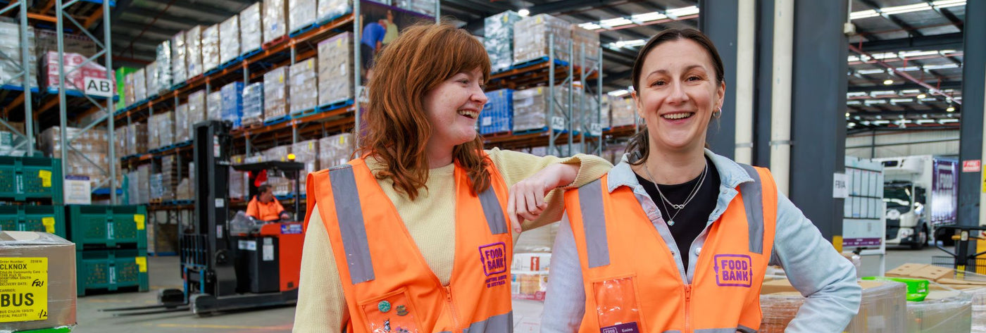 Two individuals in orange safety vests standing in a warehouse with shelves and boxes in the background.