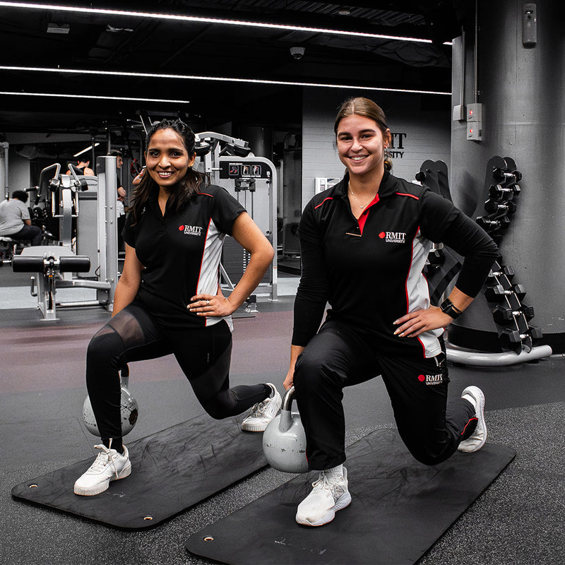 Two women doing lunges at the RMIT University gym, both are wearing black Activewear outfits consisting of leggings and tops, and they are smiling at the camera.